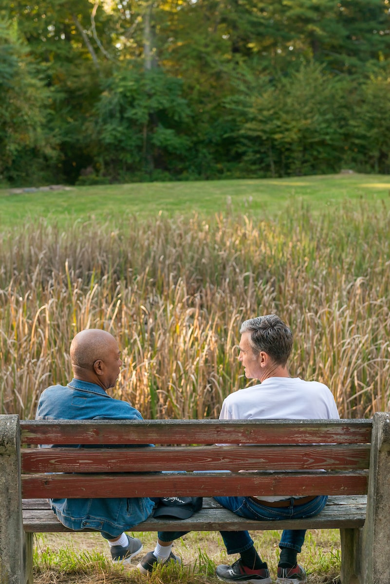 Two men talking on a wooden bench outside.