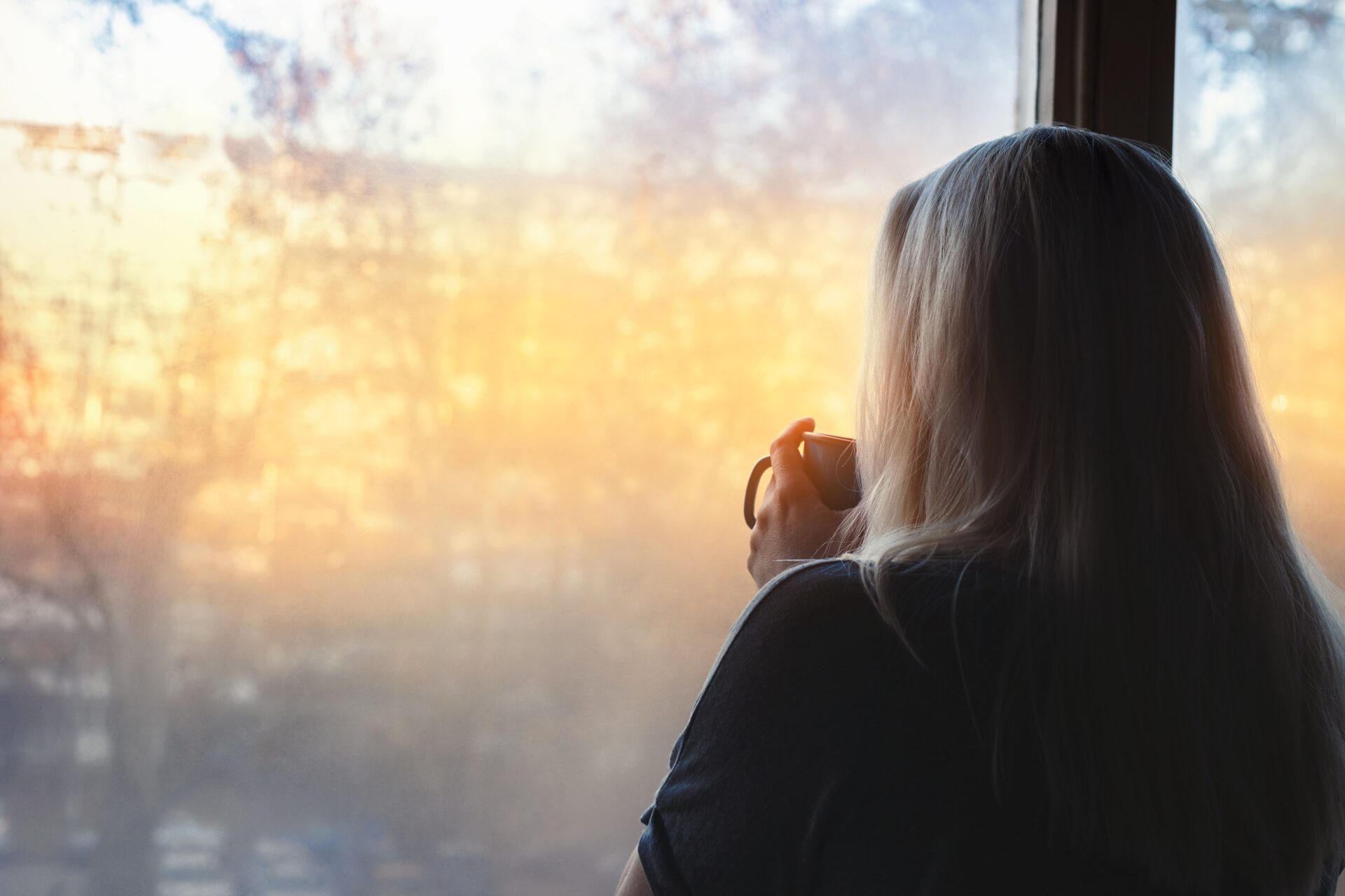 A woman looking out the window drinking coffee at sunrise.