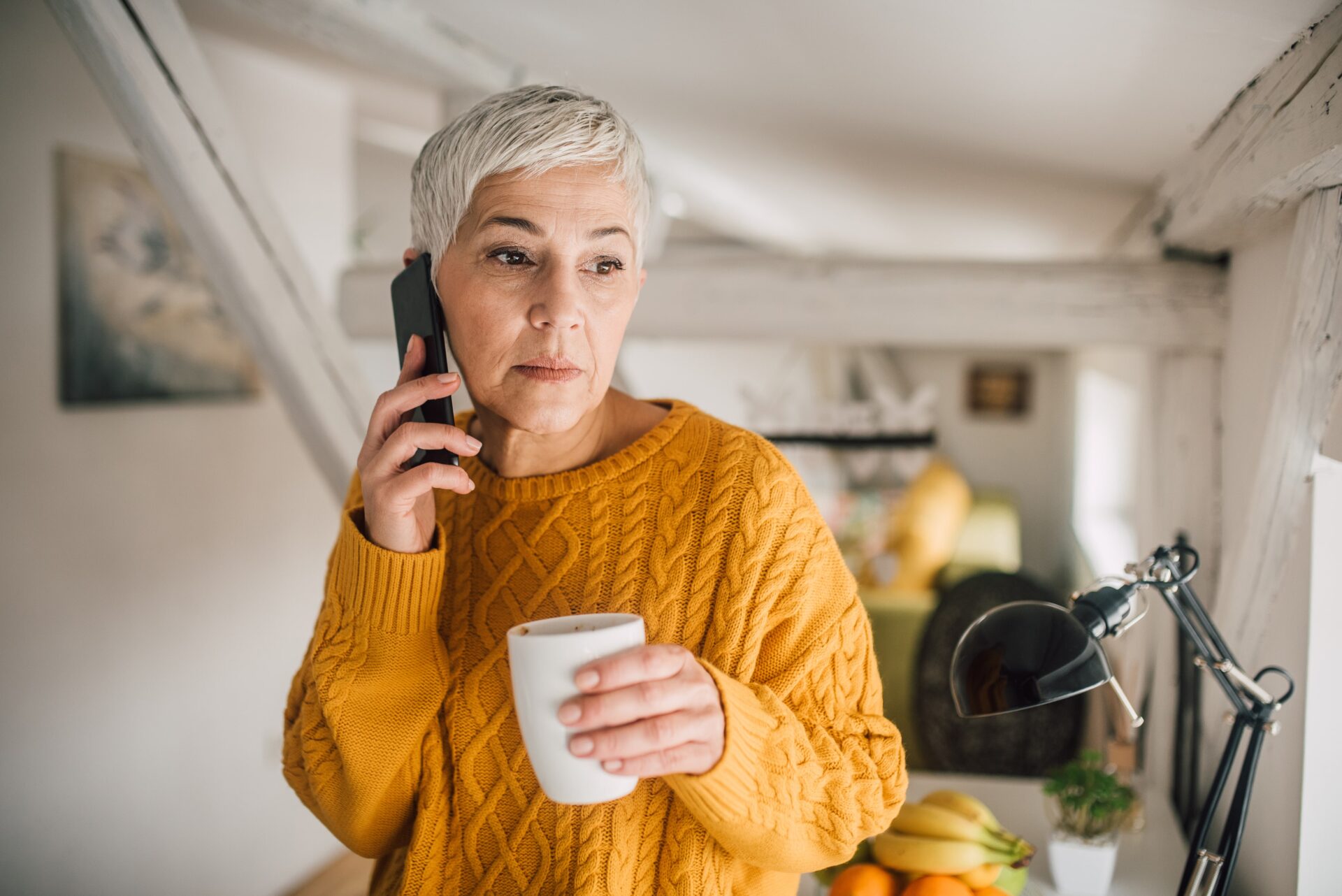 Gray-haired woman talking on the phone and drinking coffee.