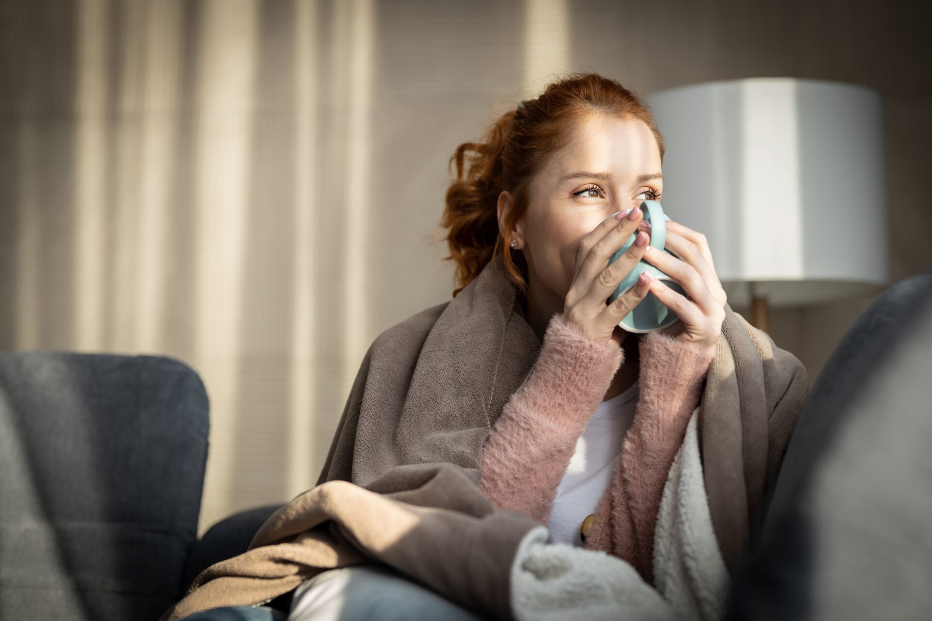 Happy woman enjoying coffee with a blanket on the couch.