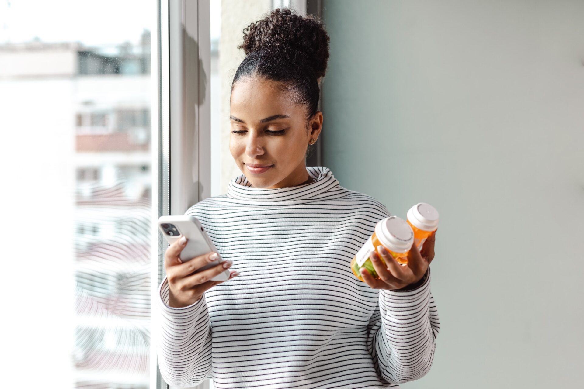 A young woman holding two medication bottles and smiling down at her smartphone.